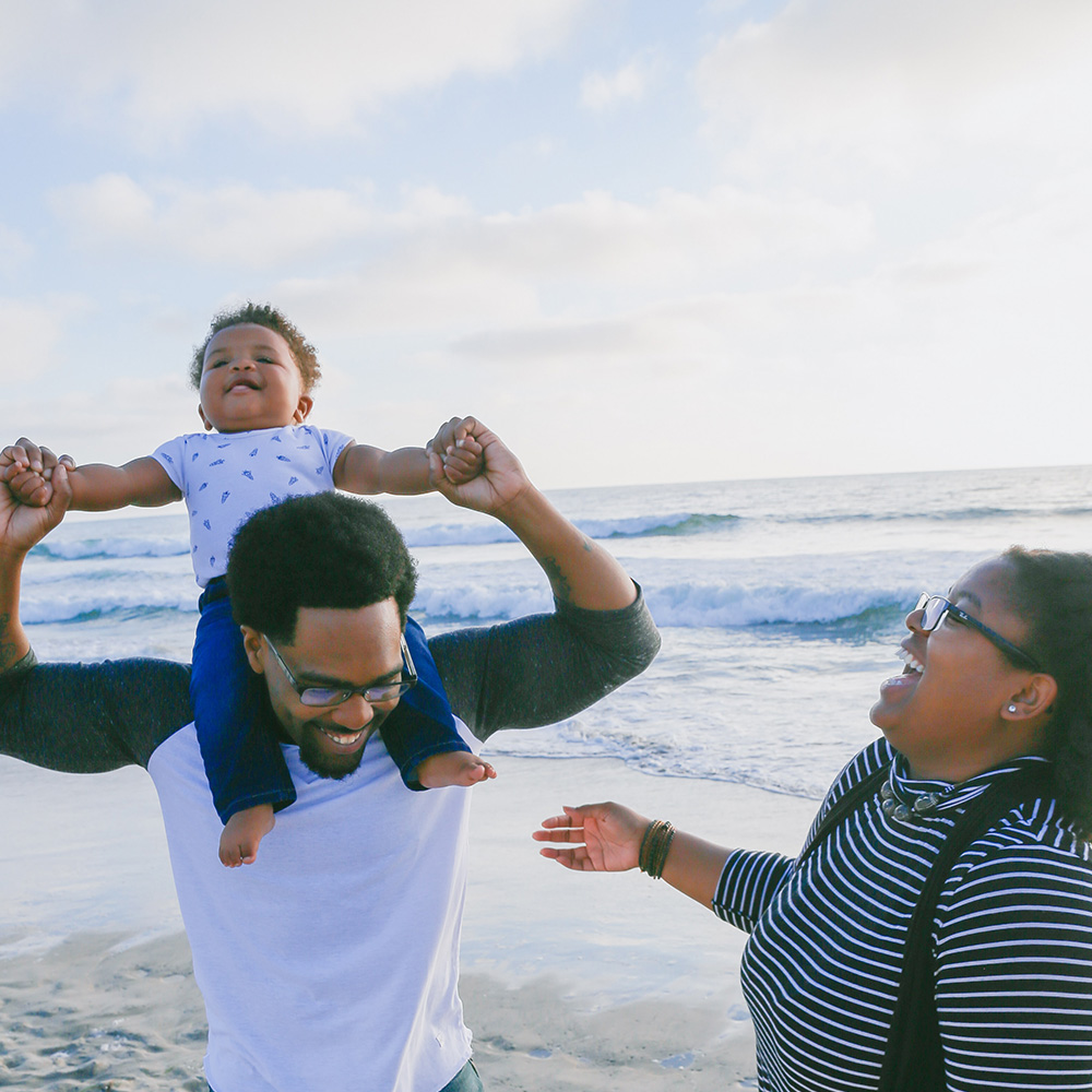 A dad has his child sitting on his shoulders while his mother laughs with joy; the family looks happy together at the shoreline of an ocean.