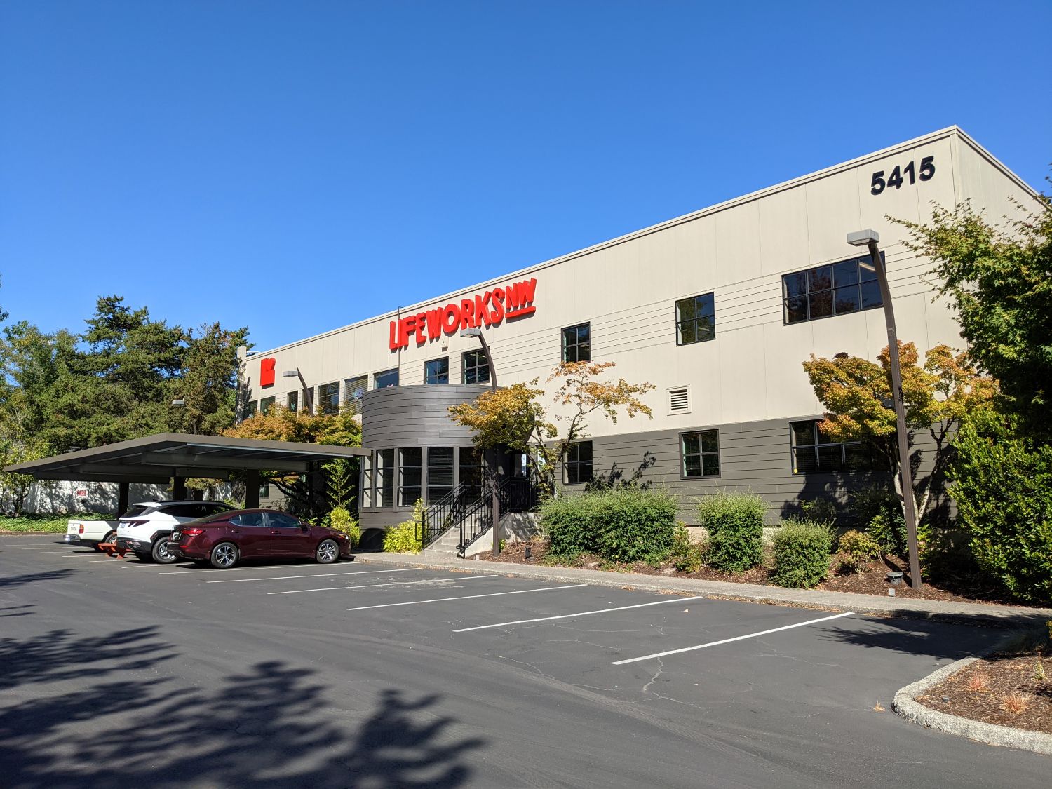 A modern two-story office building with LIFEWORKS in large red letters on the facade, located at 5415. Several cars are parked outside, and trees surround the building under a clear blue sky.
