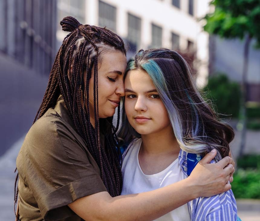 A woman with braided hair hugs a serious-looking teenage girl with long hair and blue streaks, standing outdoors in an urban setting with buildings and greenery in the background.
