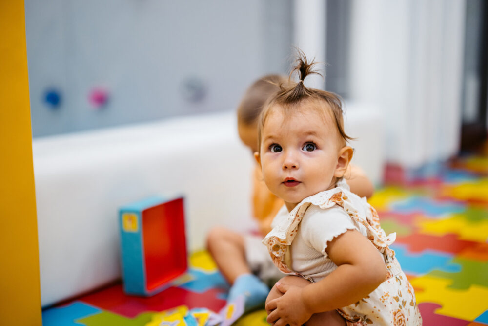 A toddler with a small ponytail sits on a colorful foam puzzle mat, looking up with curiosity. Another child is blurred in the background, playing with toys.