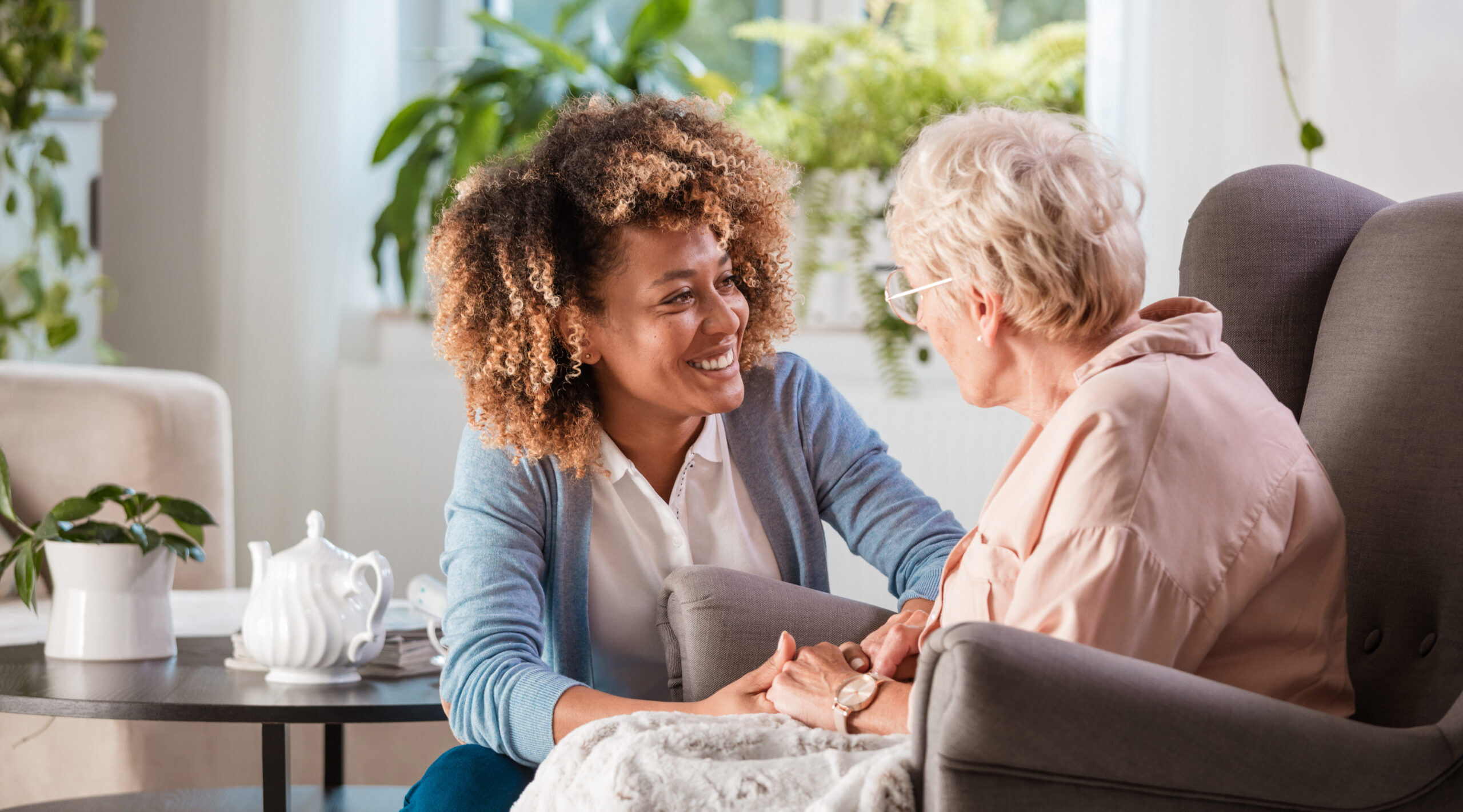 A young woman with curly hair smiles while holding hands with an older woman in glasses. They sit together in a cozy living room with plants and a tea set in the background.