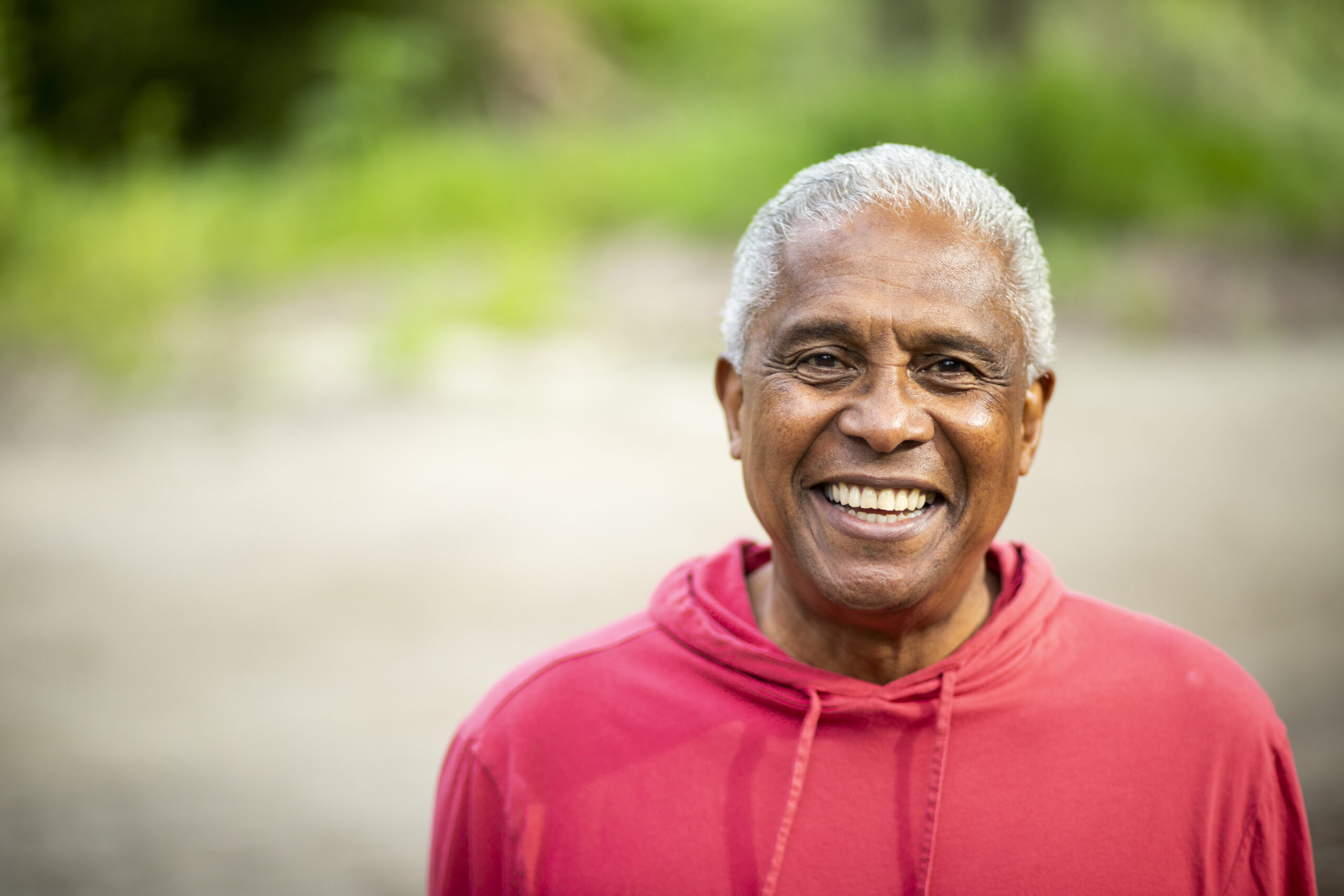 An older man with short gray hair smiles warmly at the camera while wearing a red hoodie, standing outdoors with a blurred green background.