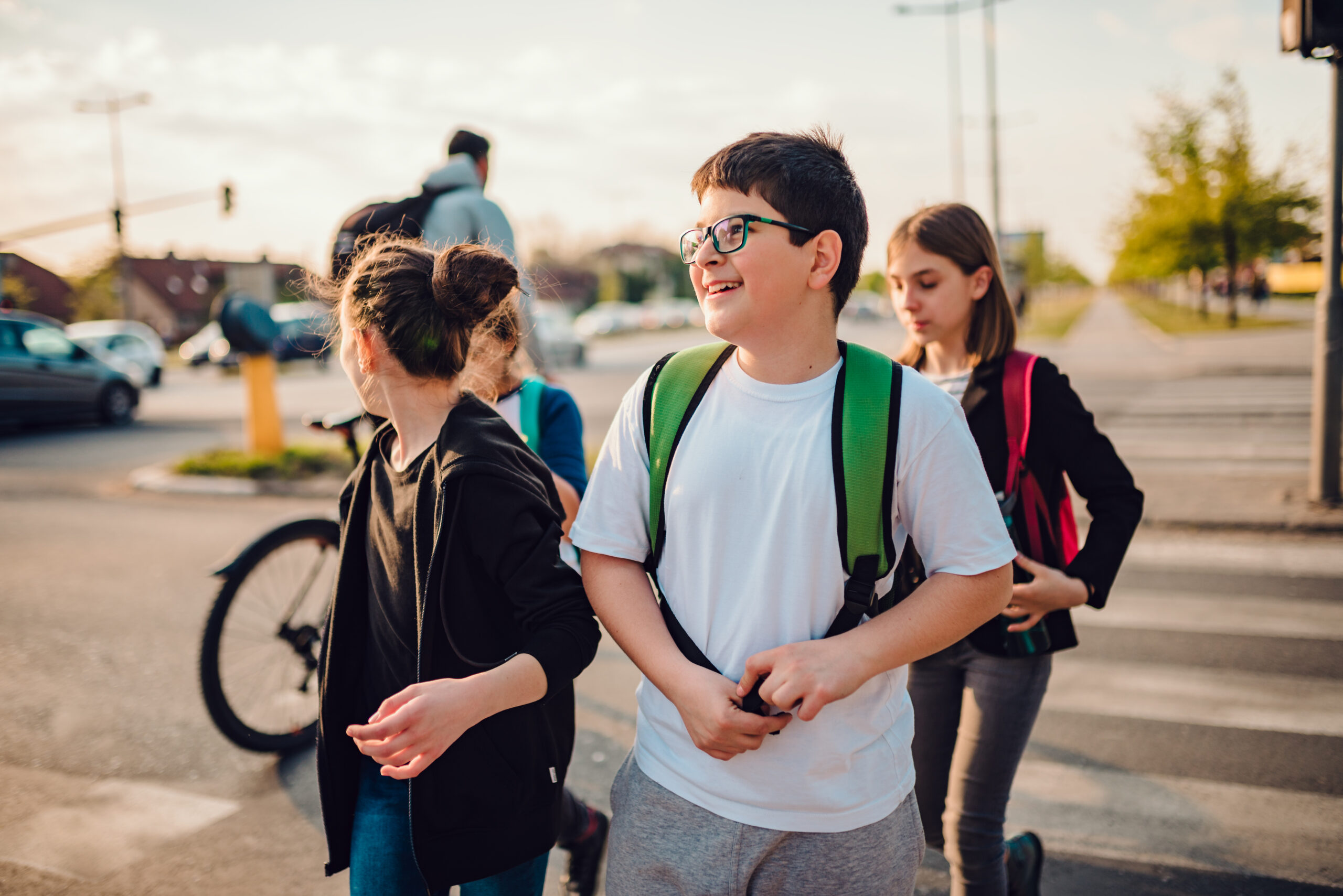 A group of children with backpacks walk together across a crosswalk on a sunny day. One child smiles while others look in different directions. A person on a bicycle is in the background.