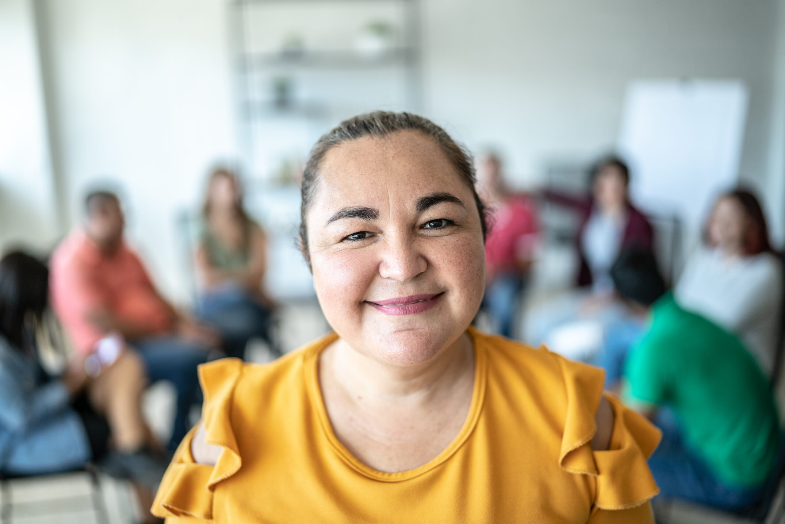 A woman wearing a yellow top smiles confidently at the camera, with a blurred group of people sitting in a circle in the background, suggesting a support or discussion group setting.