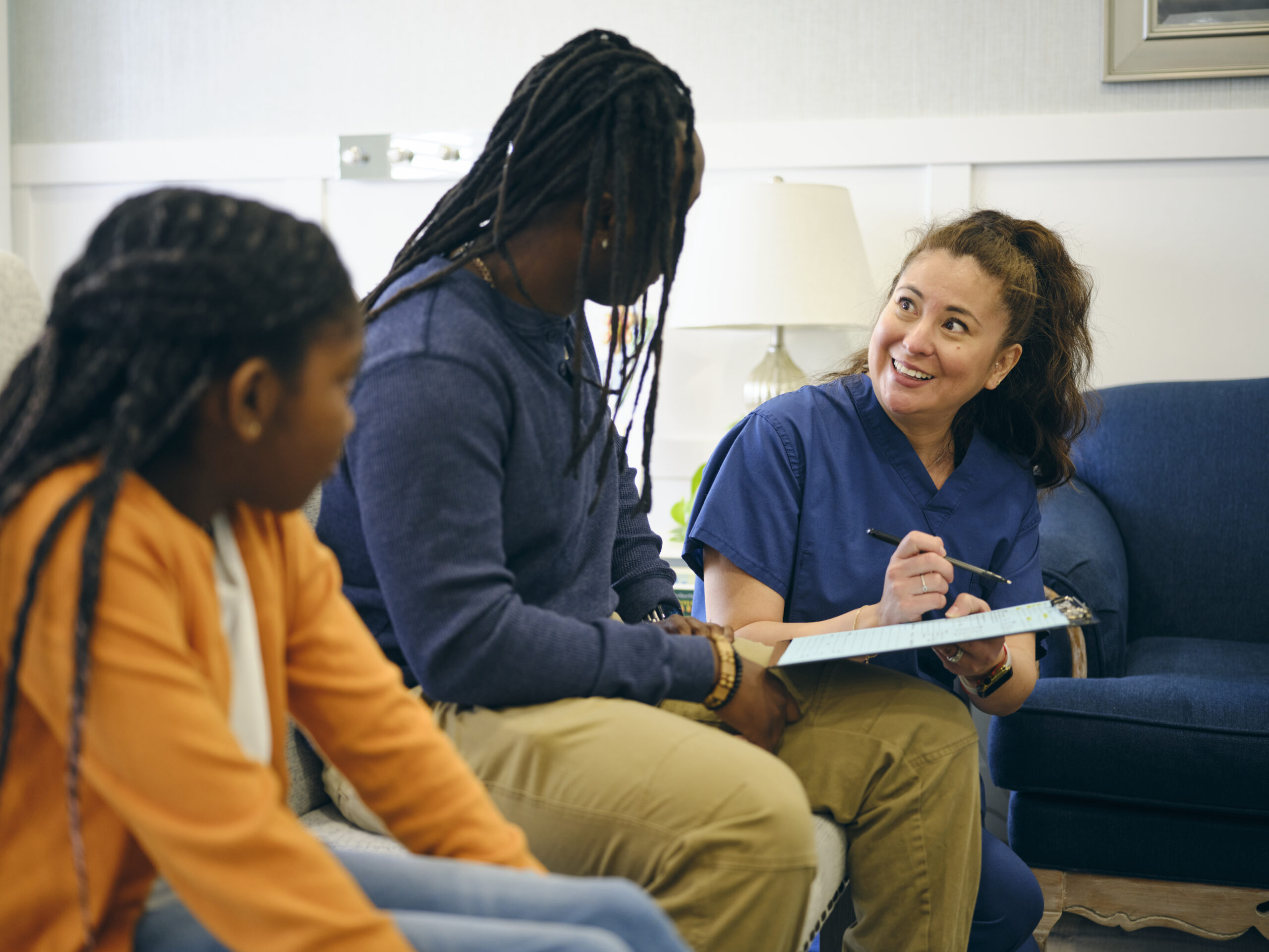 A smiling healthcare worker in blue scrubs talks with a man and a young girl, holding a clipboard and pen in a cozy, well-lit room with a navy sofa and lamp in the background.