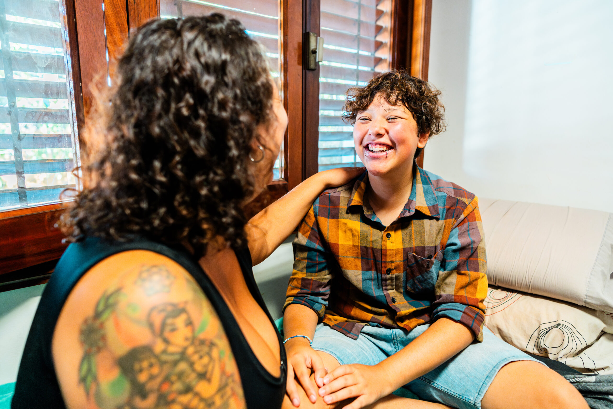 A smiling child sitting on a bed laughs while a person with curly hair and a colorful tattoo on their arm sits beside them, touching their shoulder in a brightly lit room with wooden window blinds.