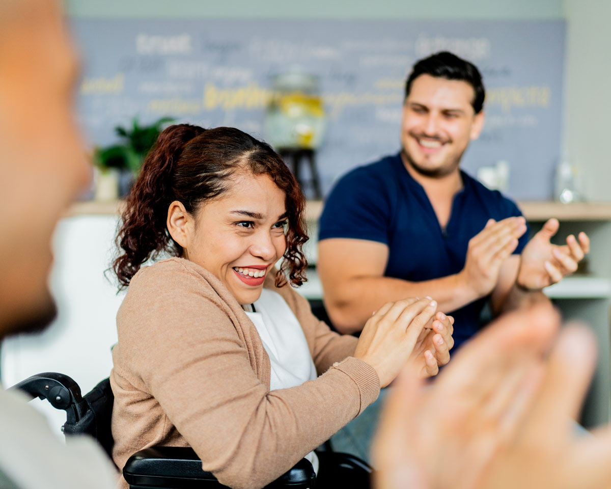 Three people sitting down, two on the outer side looking at the person in the middle with a smile while clapping their hands together. The person in the middle graciously smiles, clapping along too looking forward.
