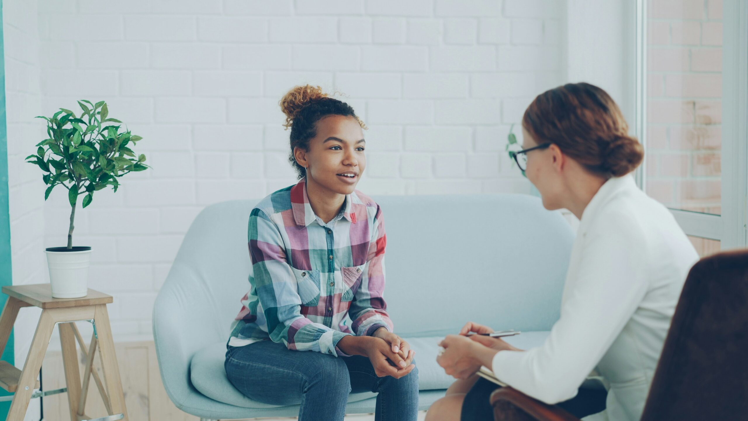 A young woman in a plaid shirt sits on a light blue couch, talking to a professional woman with glasses who is taking notes during a counseling or therapy session in a bright, modern office.