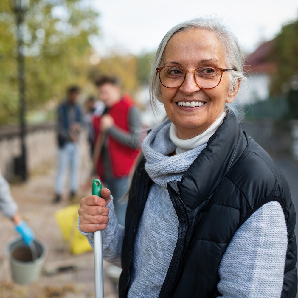 An older woman holds a shovel at a volunteer operation, where others in the background hold pails and shovels in a sandy area.