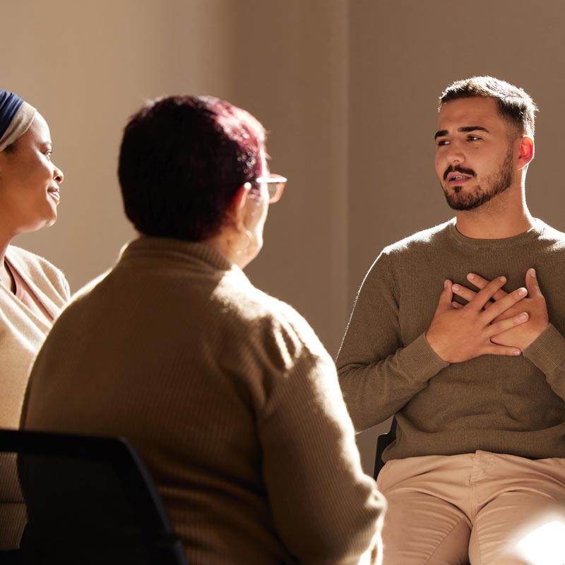 Three people sit in a support group setting, one with his hands over his heart while looking at another group member.