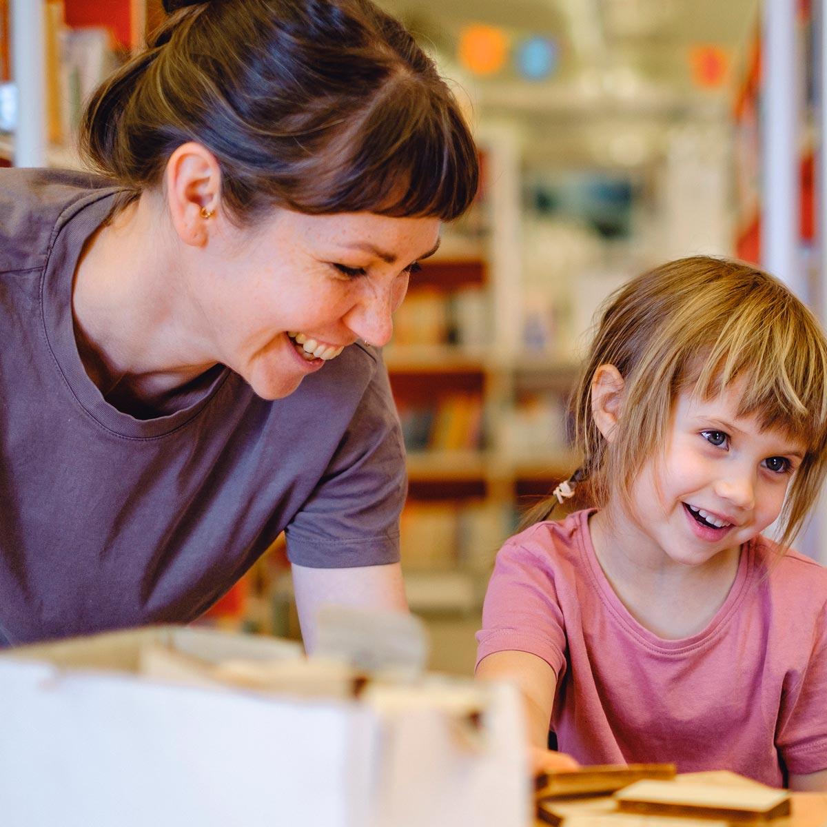 A teacher with brown hair smiles at an elementary-aged student while they work together in a classroom setting.