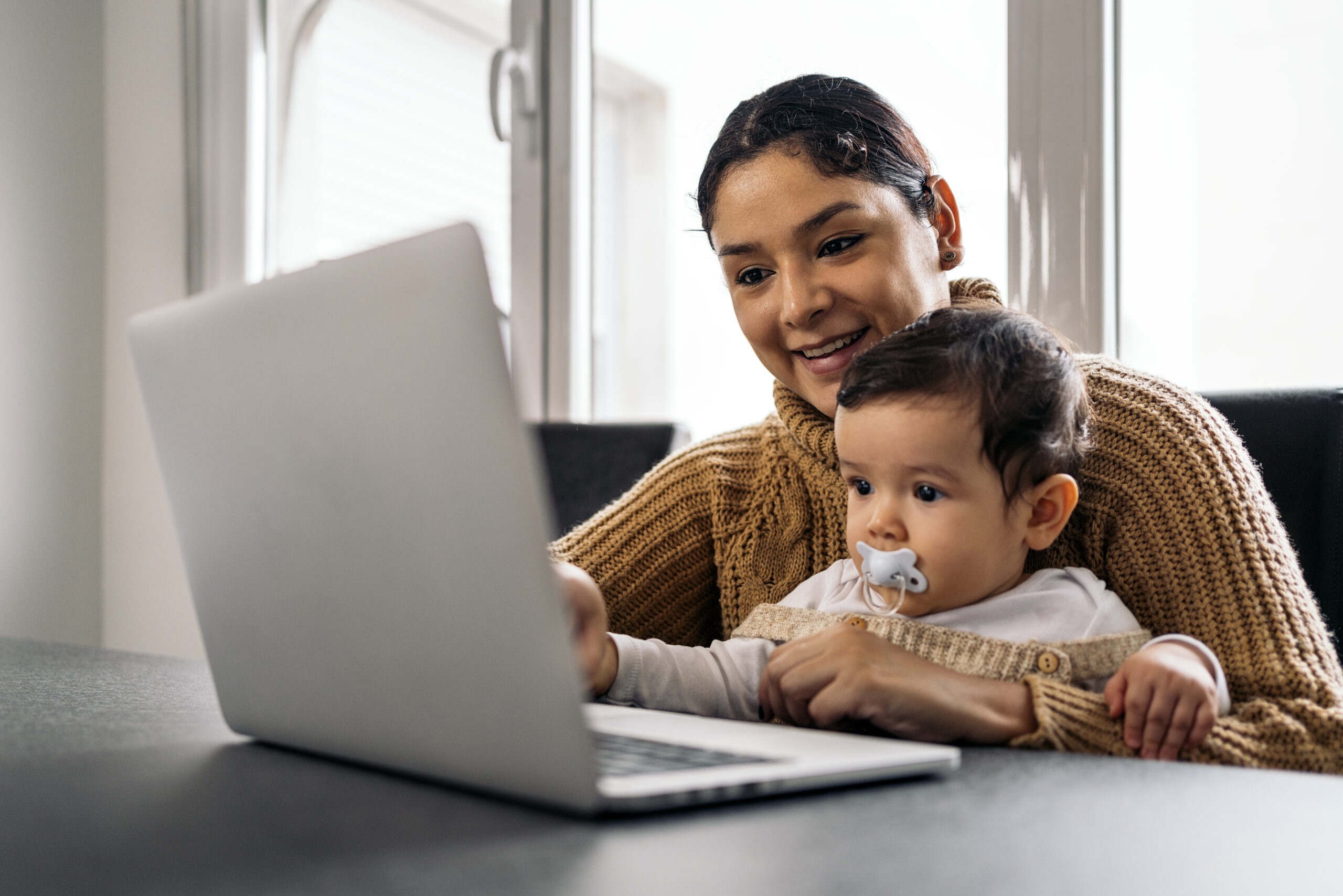 A smiling mom and her toddler sit at a table looking at a computer in front of them.