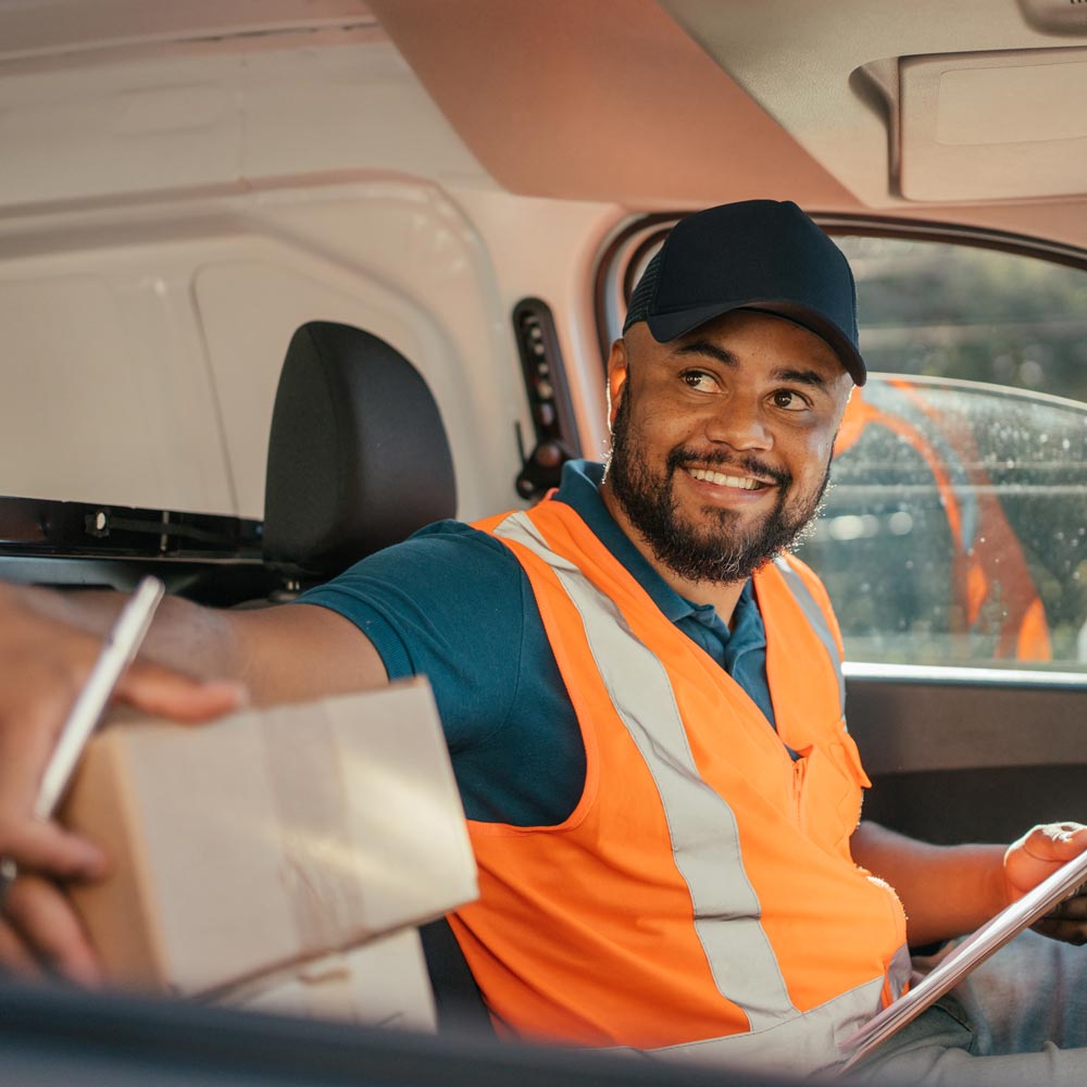 A man in a car with a fluorescent orange vest and holding a clipboard delivers packages with a smile.