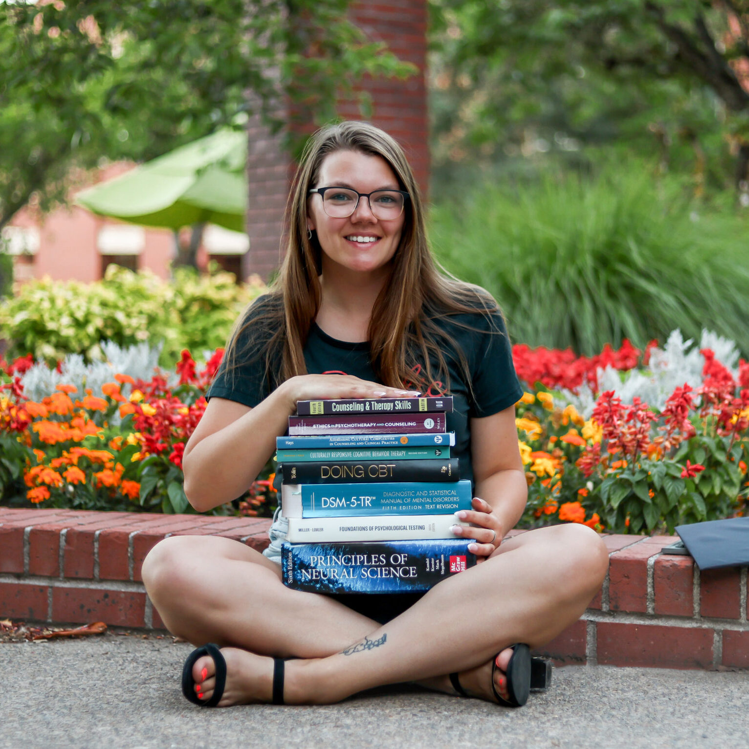 Alyssa wears glasses and is sitting on the ground with a stack of books on her lap.