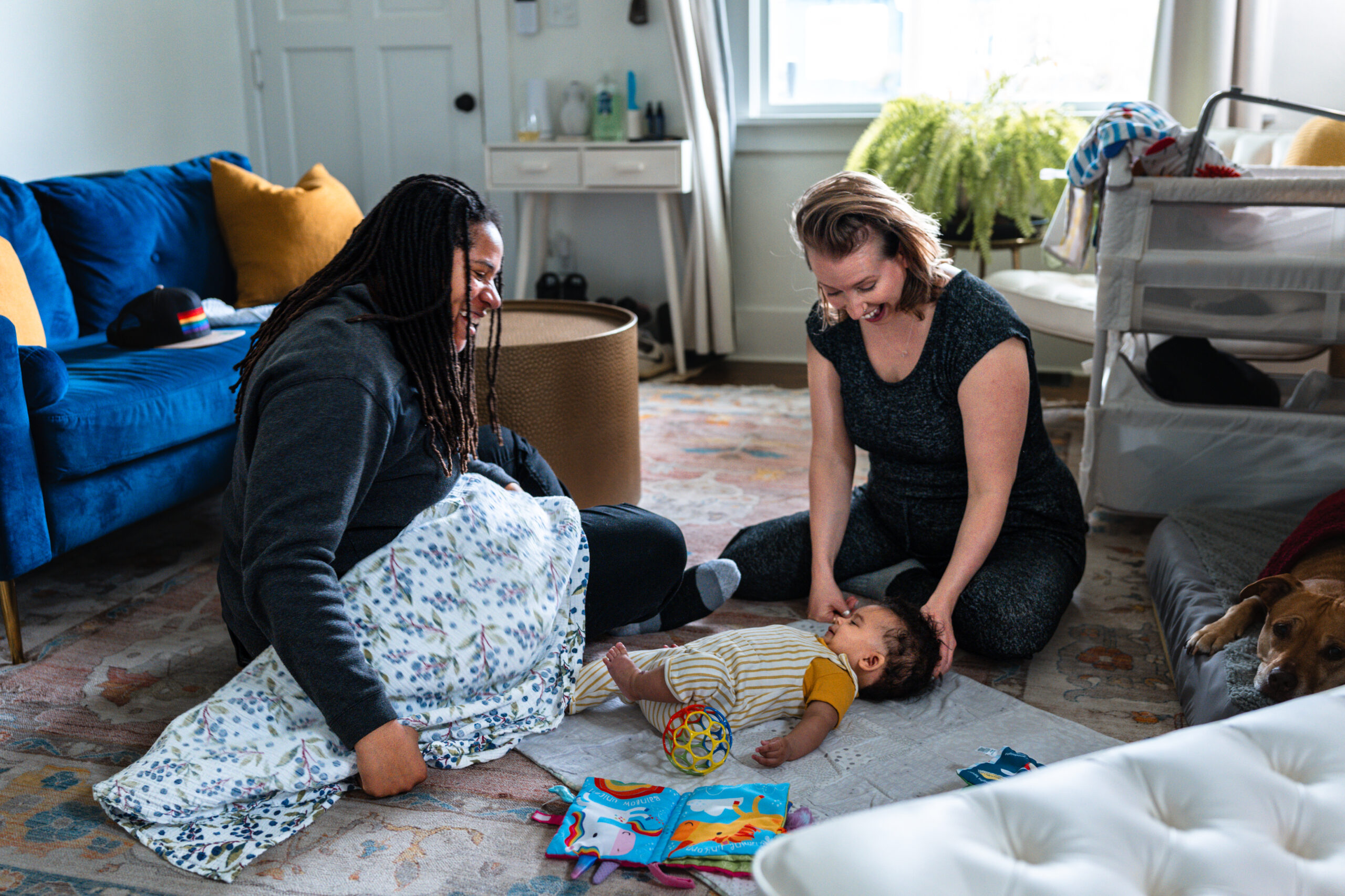 Two people sit on the floor playing with a young baby who kicks their feet happily in the air.