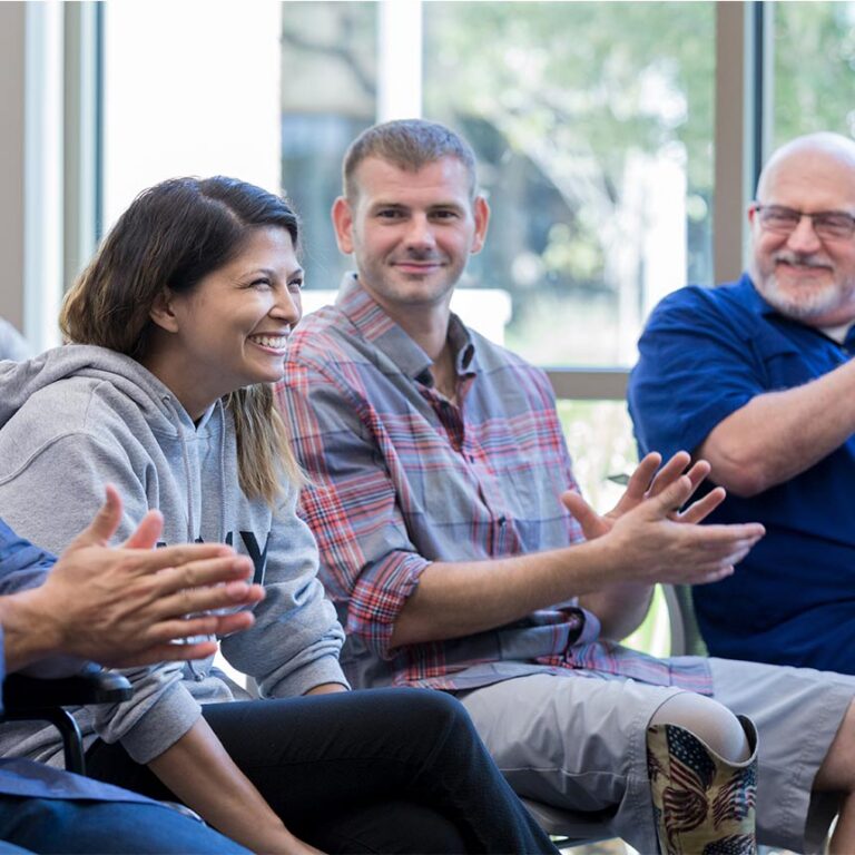 A woman sits in a group session, smiling, while others around her clap.