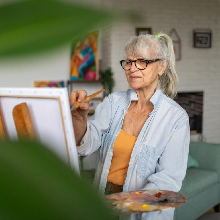 A woman with long gray hair and black glasses stands in front of an easel, painting, with a slight smile on her face.