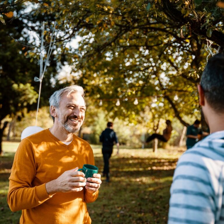 A man in his 50's in a yellow shirt holds a coffee cup in a sunny park, smiling in conversation with someone else.