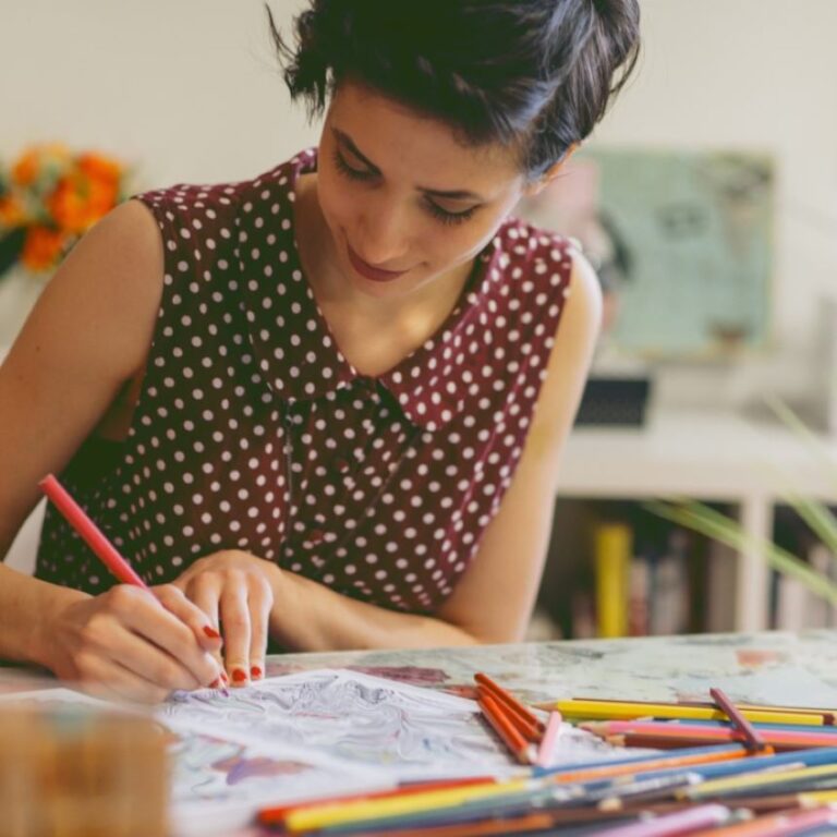 A woman in a sleeveless brown polka dot shirt colors with colored pencils and a slight smile on her face.