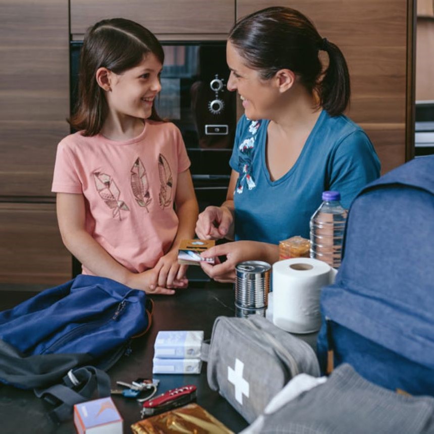 A mother and child stand in their kitchen smiling at one another, with supplies for their emergency kit sprawled out in front of them on the counter