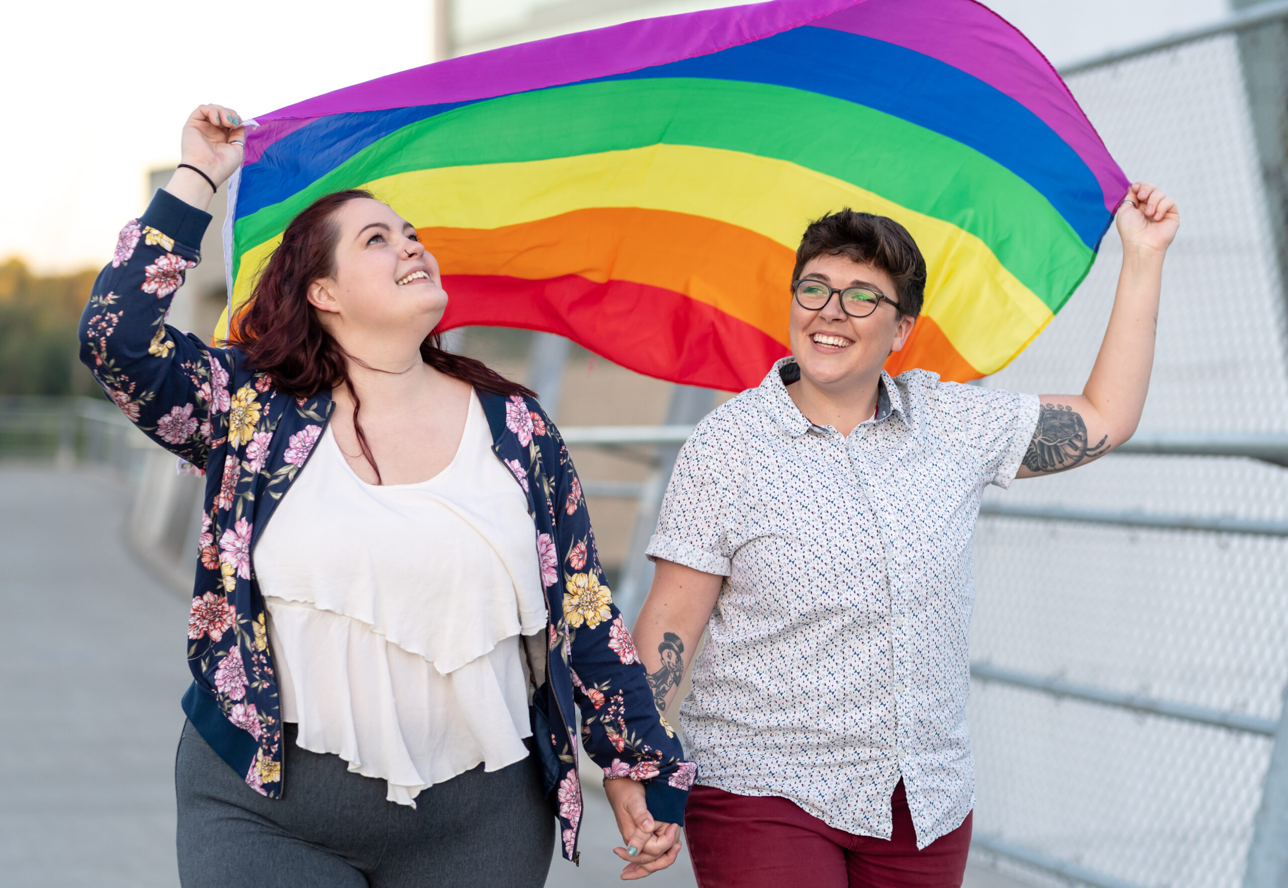 A young female couple hold hands with each other while they hold a Gay Pride Flag overhead as they walk along a Portland, OR city bridge.