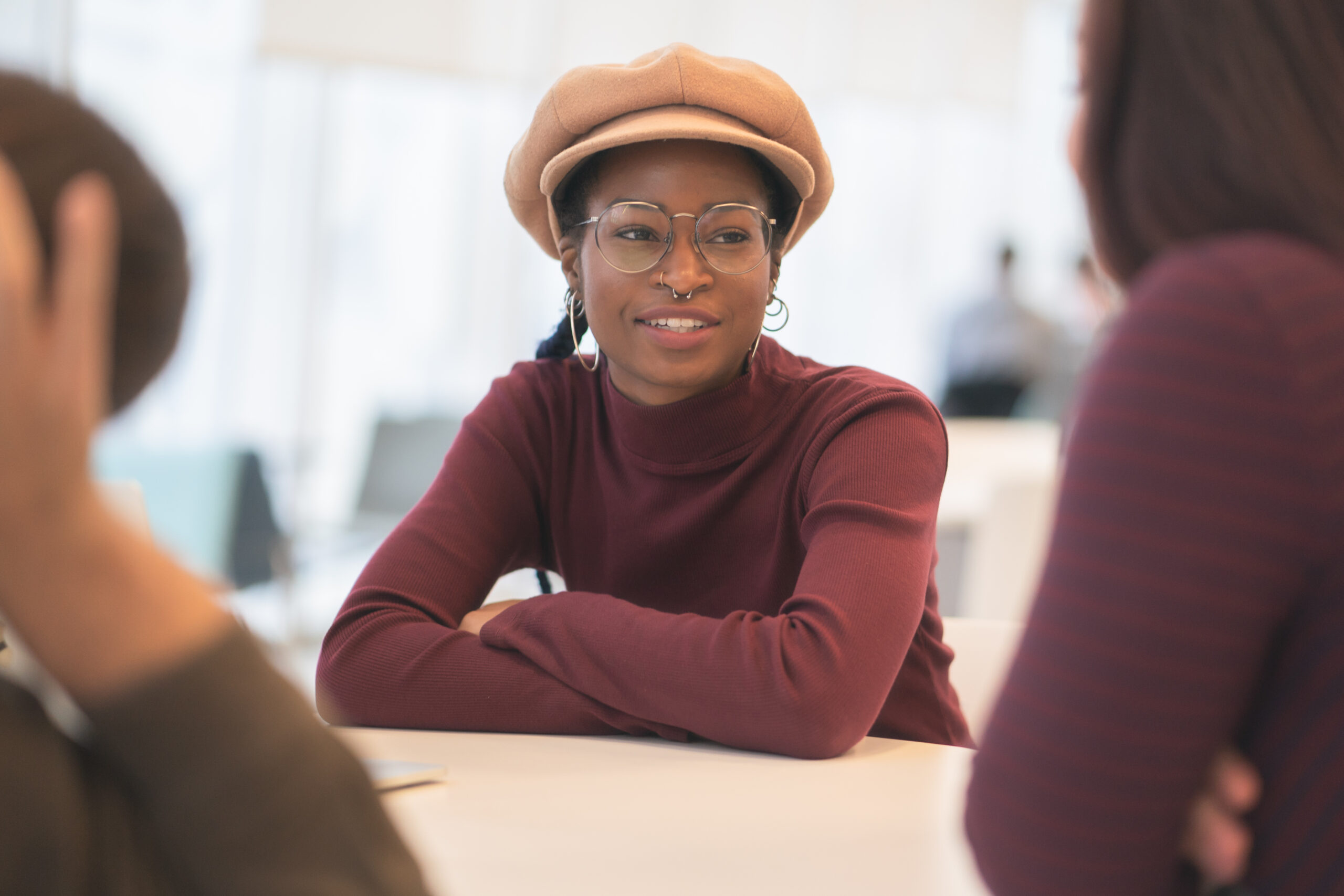 A African American woman sits among her peers. She is dressed casually, is wearing a hat and glasses. She is smiling as she listens to her peers input.