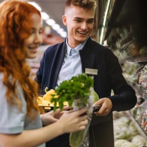 A young grocery story employee helps a customer select a product, both smiling.