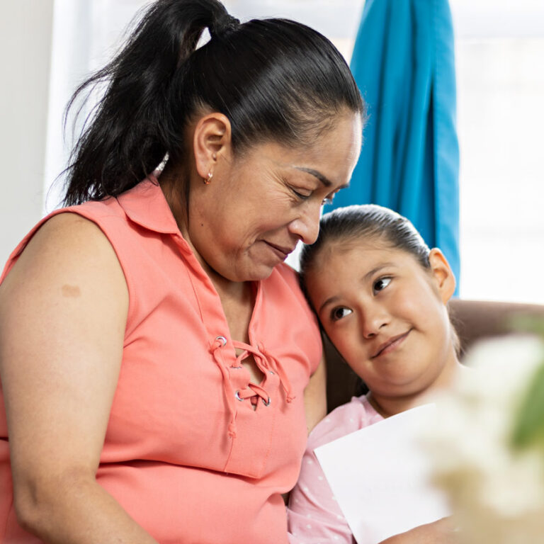 A young child looks up with a gentle smile toward an older woman, sitting together on a couch.