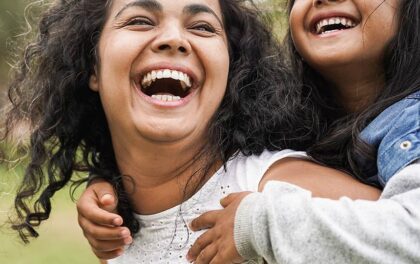 A mom walks happily through a park with her daughter on her back, both smiling.