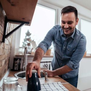 A man stands in his kitchen, washing dishes with a smile on his face.