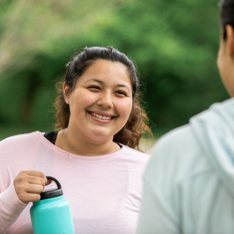 A woman in a pink sweatshirts holds a water bottle and smiles at someone in conversation.