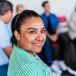 A women in a group of people sitting in chairs in a circle looks back toward the camera with a bright smile.