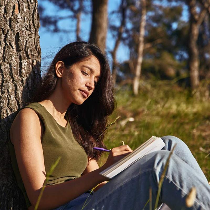 stressAwareness_tree_journal A woman sits calmly with her back on a tree trunk, writing in a journal.