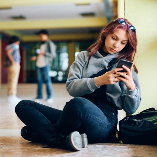 A teenager sits on the floor of her school looking at her cellphone.
