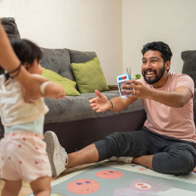 A dad sits on the ground and celebrates, smiling, as his toddler child walks toward him in their living room.