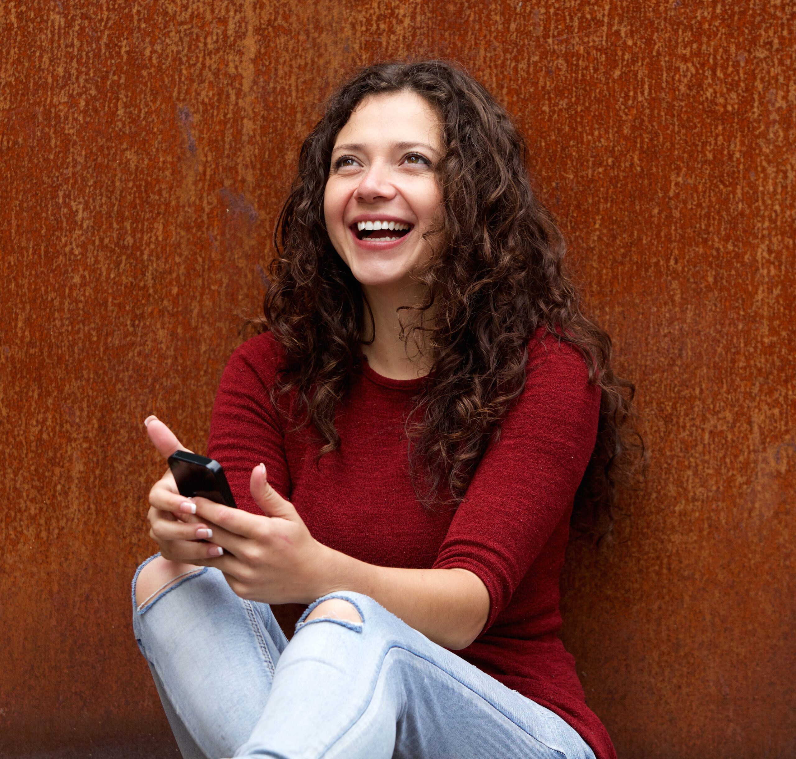 Portrait of smiling young woman sitting against brown wall with mobile phone